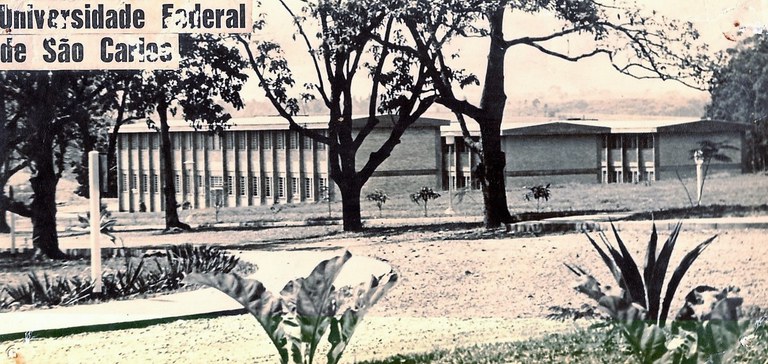 Fotografia antiga, aparentando estar desgastada pelo tempo e em preto e banco. Mas está mais amarelada, devido ao envelhecimento. Como conteúdo principal aparecem três prédios, cada um com andar térreo e um primeiro andar. Possuem cerca de 30 janelas, cada um deles. Isso considerando a visão em ângulo obliquo, utilizada.  Antes dos prédios, vemos 5 árvores grandes e alguma poucas mudas de plantas. Além disso, existe um poste branco, ao lado de uma calçada curvilínea. Ao fundo da foto existe uma vegetação, podendo ser uma fazenda ou uma floresta, ou ambos.