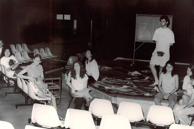 Fotografia em preto e branco ambientada em um teatro pequeno e cujo palco é semi hexagonal, na frente. O palco está elevado cerca de 70 centímetros do piso. Na plateia encontram-se cadeiras de assento plástico, fixas ao chão. Sobre o palco encontram-se alguns papeis e uma lousa. Na foto temos 7 mulheres jovens sentadas, nas cadeiras ou no palco, e um rapaz com pochete, de pé. 